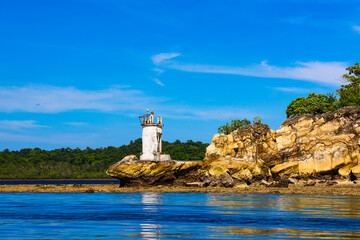 A picturesque lighthouse stands on a rocky coast, surrounded by blue water and a dense forest under...