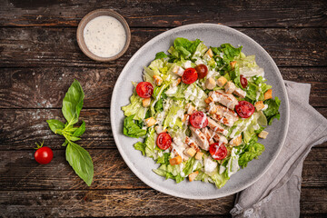 Fresh caesar salad with chicken breast, croutons, lettuce, parmesan cheese, sauce and tomatoes on wooden background, top view. High quality photo