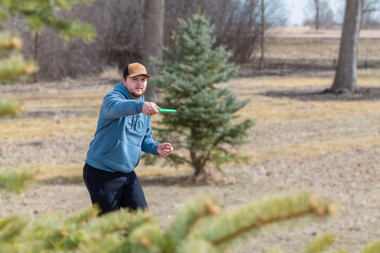 Man Throwing Green Disc Golf Disc with Foreground Tree Branches in Early Spring - Blurry Background