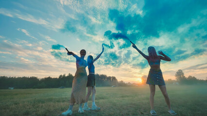 Three school friends enjoying a vibrant celebration, waving blue smoke bombs against a picturesque...