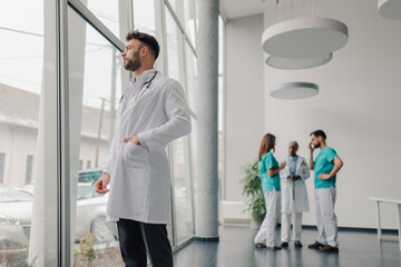 Doctor looking out window while medical team discussing in hospital lobby