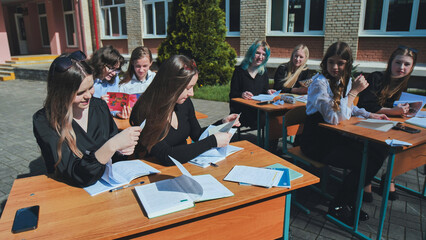 High school girls studying and reading together at desks in a lively schoolyard on a beautiful, sunny day