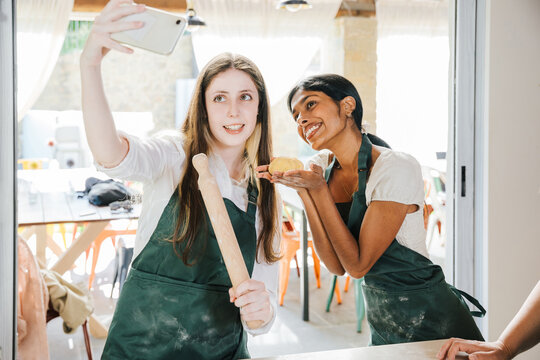 Two young friends take a photo with smartphones at a fresh pasta cooking class - show off their freshly made dough and smile at the phone while wearing work aprons