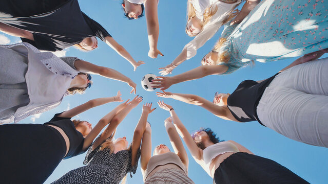 High school students standing in a circle with their hands raised towards a soccer ball under a clear blue summer sky