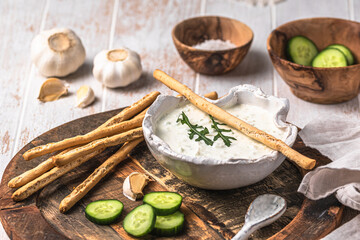 Greek tzatziki in a handmade white bowl with ingredients and cracker sticks on a wooden background