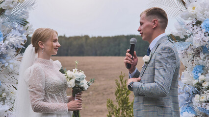 Groom speaking at wedding ceremony with microphone, holding vows and looking at bride with love,...