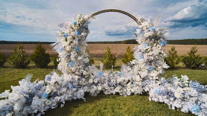 Elegant wedding arch decorated with white and pale blue blooms, standing amid grassy landscape...