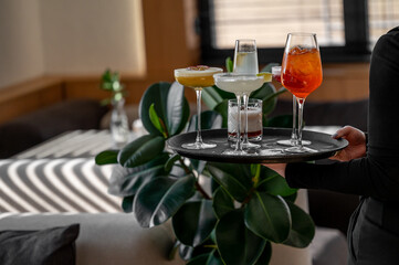 A waiter holding a tray with assorted cocktails, including margarita, Aperol spritz, whiskey sour, and champagne, in a chic restaurant interior with modern decor.