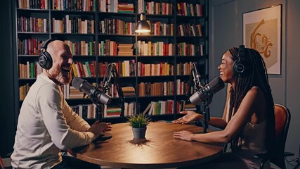 A video of two people recording a podcast in a cozy library setting. Side angle captures their conversation, microphones, and bookshelves in the background.