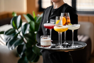 A waiter in a black uniform holds a tray with six colorful cocktails and a white drink, served in various glassware, with a cozy interior and green plants in the background.