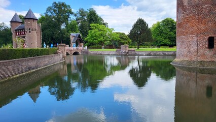 Obraz premium A historic castle stands near a calm lake, reflecting lush trees and a bright blue sky. Visitors enjoy the serene atmosphere in this picturesque location.