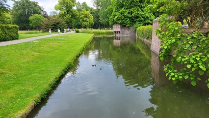 Obraz premium Visitors enjoy a tranquil afternoon in a lush park, with manicured lawns reflecting serene waters and elegant statues lining the pathways under a partly cloudy sky.