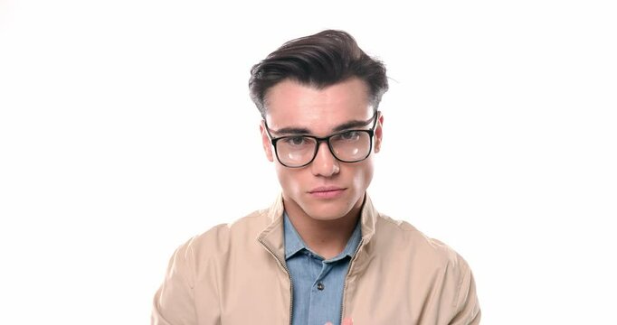 portrait video of casual young man with glasses rubbing palms, adjusting beige jacket and looking to side in front of white background