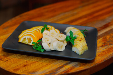 Beautifully chopped fruits on a black plate decorated with mint, all this stands on a wooden table in a restaurant