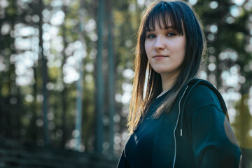 Fototapeta premium A woman in a black top and light jeans stands confidently in a sunlit forest, with soft lens flare and tall trees creating a dramatic and empowering atmosphere.