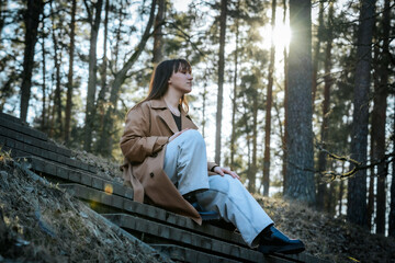 A woman in a trench coat and light jeans sits thoughtfully on forest steps in soft sunlight, surrounded by tall trees with a subtle lens flare.