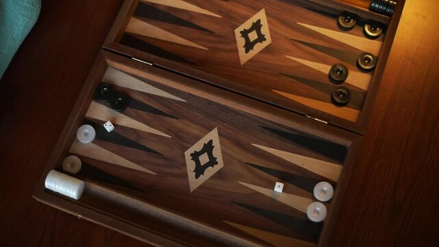 A close-up of a backgammon game in progress, with a hand moving a white piece over the wooden board. Black and white pieces are arranged, and the moment captures focused strategic play.