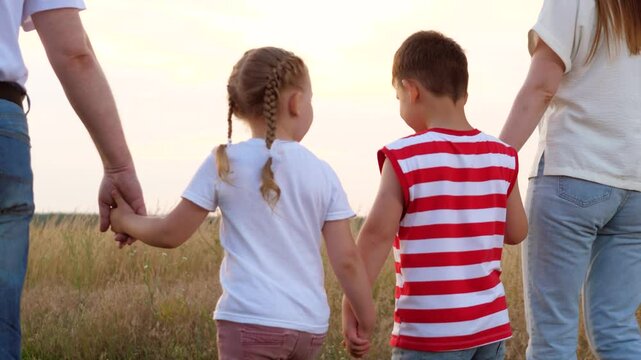 Happy little children walk joining hands with parents across summer meadow at sunset backside view. Loving children son and daughter discover nature with parents. Family and children walk in field