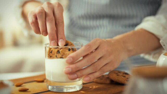 Hands dip a chocolate chip cookie into a glass of milk at a wooden table. The inviting setting, filled with light, suggests a warm afternoon treat during a relaxing break