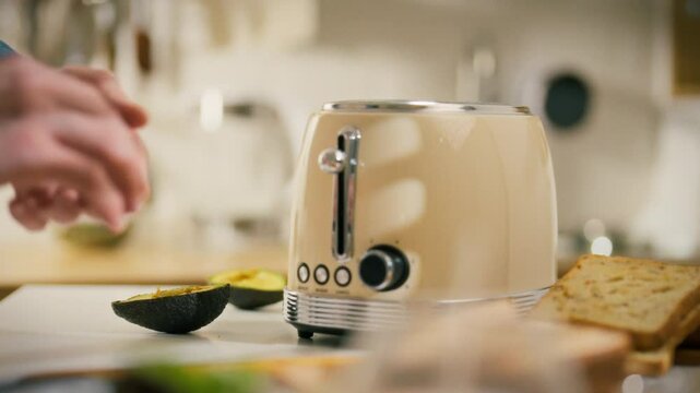 A person places a slice of bread into a beige toaster in a warm, inviting kitchen. Fresh avocado and additional bread sit nearby, showing preparation for a healthy breakfast