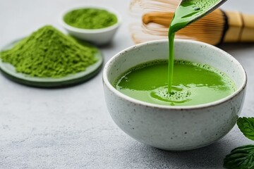 Ceramic bowl of freshly stirred matcha tea with bamboo whisk and powder on light gray surface