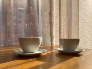 Two minimalist white coffee cups casting long shadows on a wooden surface, with sheer curtains and city buildings in soft focus in the background