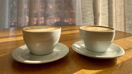 Two cappuccino cups with beautiful latte art, bathed in warm, morning sunlight streaming through sheer curtains with city buildings in soft focus behind