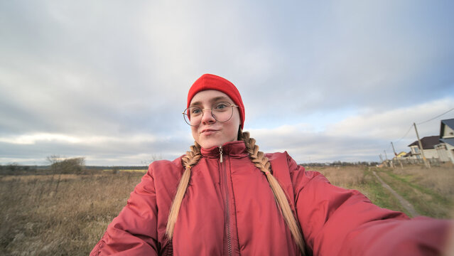 Young girl with braided hair, glasses, red jacket capturing selfie amid rural landscape, showcasing casual outdoor style