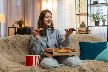 Happy young Caucasian woman eating delicious junk food pizza and popcorn sitting on sofa couch...