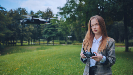 Young woman piloting a drone in a green park using a remote controller, enjoying a sunny day...