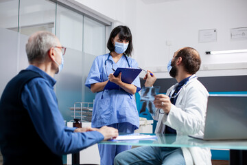 Old man sitting in clinic and listening to young healthcare workers discuss his lung radiograph and treatment options. Doctor speaking to nurse with clipboard as senior patient awaits xray explanation