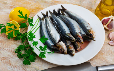 Plate of raw sardines on wooden table with sprigs of fresh fragrant parsley, garlic cloves, lemon and oil. Main ingredients for cooking