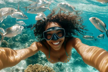 Fototapeta premium A stunning and sexy happy black young woman with curly hair in a swimming mask and fins snorkeling among the corals. Sunlit underwater beauty. A dense school of silver fish swims next to her.