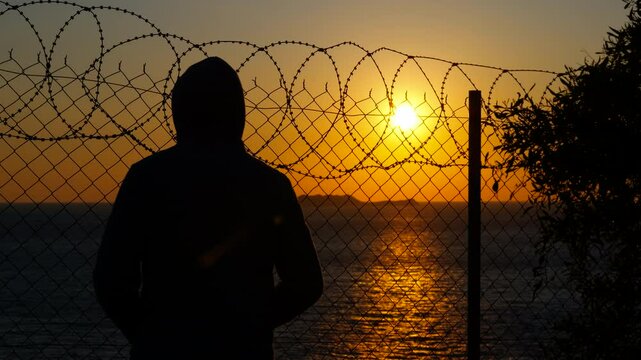 Refugee at the border at sunset. Silhouette of a hooded young man and a young man with a baseball cap, standing behind a barbed wire fence, contemplating a beautiful sunset over the sea