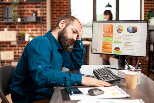 Tired businessman sitting at desk and holding his forehead in startup office. Exhausted male employee having stacks of financial paperwork and computer on table, reflecting demanding work schedule.