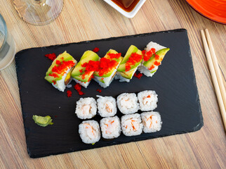 Sushi set of makizushi rolls filled with boiled prawn and colorful uramaki with raw salmon, mango and cucumbers topped with thin avocado slices and masago roe served with soy sauce and wasabi