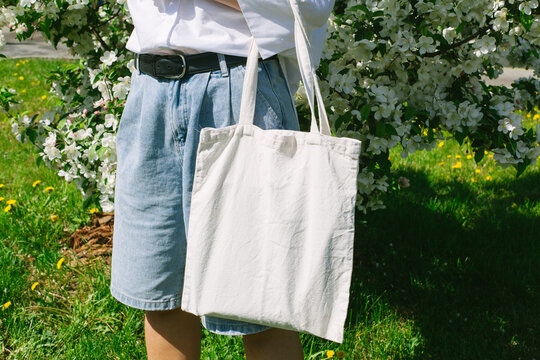 Woman hand holding eco bag. Female hold white canvas textille mesh zero waste shopper on city street, green apple tree flowers background. Blank mock up tote bag, place for text design logo.