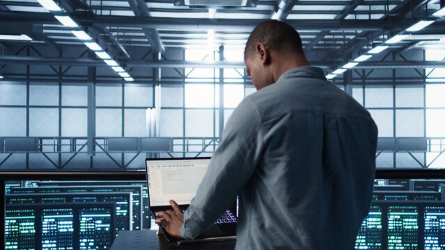 Computer scientist in server room coding on laptop, developing disaster recovery plans. African american man in data center testing failover scenarios using notebook, camera B - Powered by Adobe