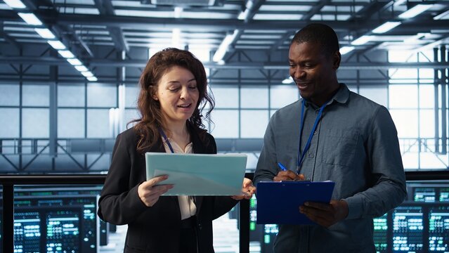 Data center management executives reading paperwork files, reviewing system diagnostics. Server farm colleagues looking over maintenance schedules documents and cybersecurity protocols, camera B