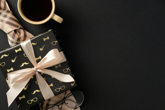 Gift box with beige ribbon, coffee cup, plaid tie, and glasses on a black background. Perfect for Father's Day, men birthdays greetings concept