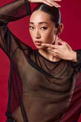 Asian woman in sheer top posing on red background with hands on head and face