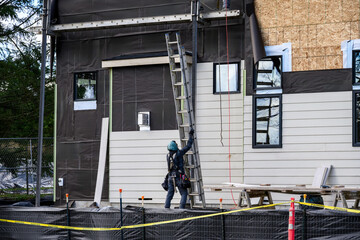 Construction worker carrying an aluminum extension ladder on home construction job site, moisture resistant membrane paper and wood siding being installed
