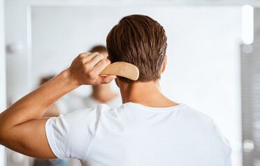 Male Haircare. Back view of unrecognizable young guy brushing his glossy hair with wooden comb,...