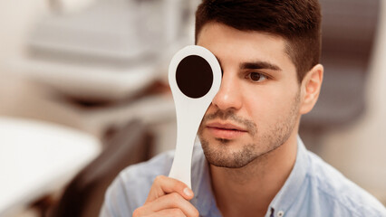 A young man focuses intently while holding an occluder over one eye during a vision examination in a modern eye care clinic, highlighting the importance of visual health.