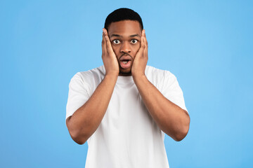 Fototapeta premium Overwhelmed Black Man Cupping Face In Hands Looking At Camera In Shock Standing On Blue Background. Studio Shot