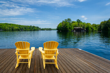 Two yellow Adirondack chairs on a wooden dock greet the tranquil Muskoka summer morning, facing the...