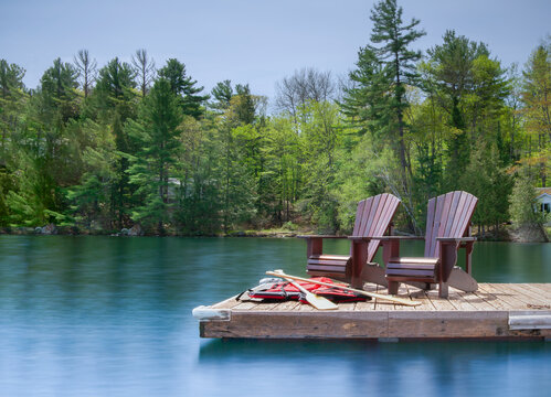 Two Muskoka chairs rest on a wooden dock overlooking a calm lake, captured in a long exposure. A white cottage stands behind them, with life jackets and canoe paddles scattered nearby.