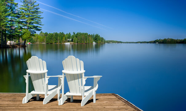 Two white Adirondack chairs on a wooden dock face the calm Muskoka lake, its glassy surface from a long exposure reflecting the sky. Cottages among green trees add to the serene scene.