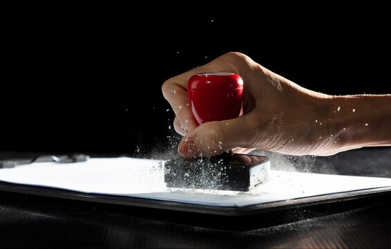 Against their will, hand delivers stamp, marking contract with forced approval. Ink signs though carrying authority, lack genuine force. Office air is thick with tension as document is pushed through