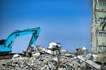 A teal excavator demolishes a building, sitting on a pile of rubble against a clear blue sky.